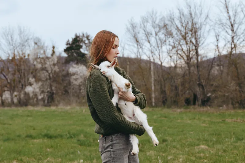 Woman outdoors embracing a goat kid in a field, calm, affectionate moment amid greenery and nature, soft light, gentle breeze, rustic countryside backdrop, lifestyle scene