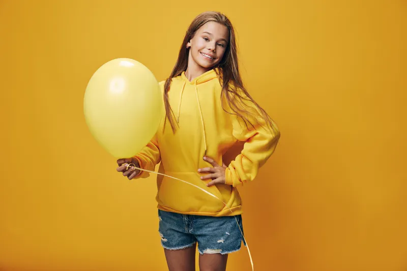 Girl in yellow hoodie holding balloon and smiling on yellow background wearing denim shorts standing with hand on hip in studio light.
