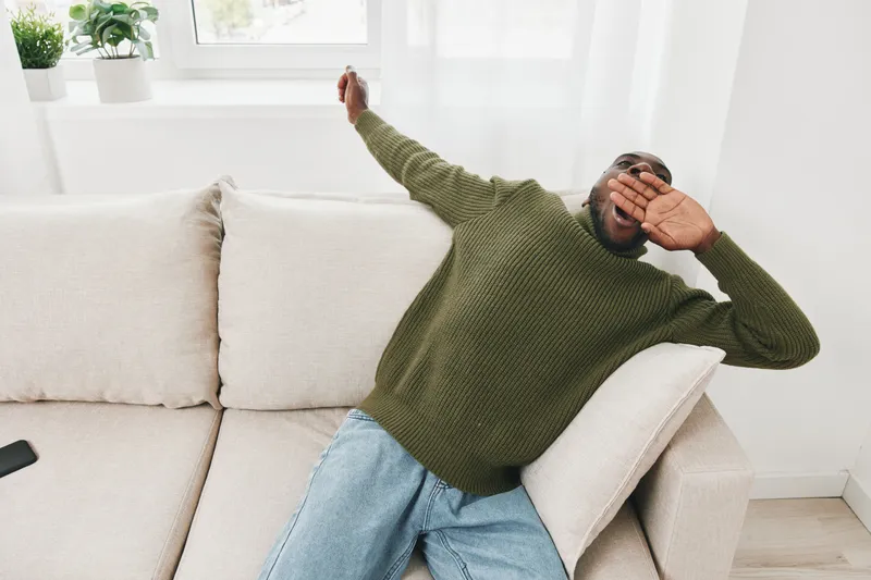 Relaxed young African man in green sweater yawning on a sofa in a bright living room, depicting comfort and leisure
