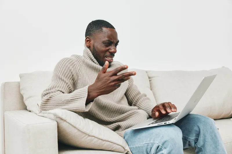 Young African man in a cozy sweater showing frustration while working on a laptop at home, capturing the essence of remote work challenges
