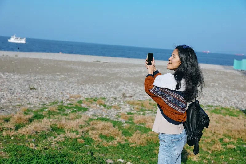 Beach scene with a woman on the shore, capturing a moment with a smartphone. Casual cardigan, backpack, and coastal landscape under blue sky, conveying relaxed travel and outdoor photography