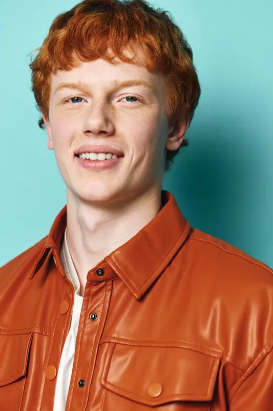 Young man with red hair and pale skin wearing brown leather jacket smiling with teeth on blue background in studio portrait, casual style and confident expression.