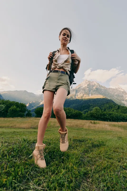 Young woman with backpack hikes through a grassy field with mountains in the background enjoying outdoor adventure and nature exploration.
