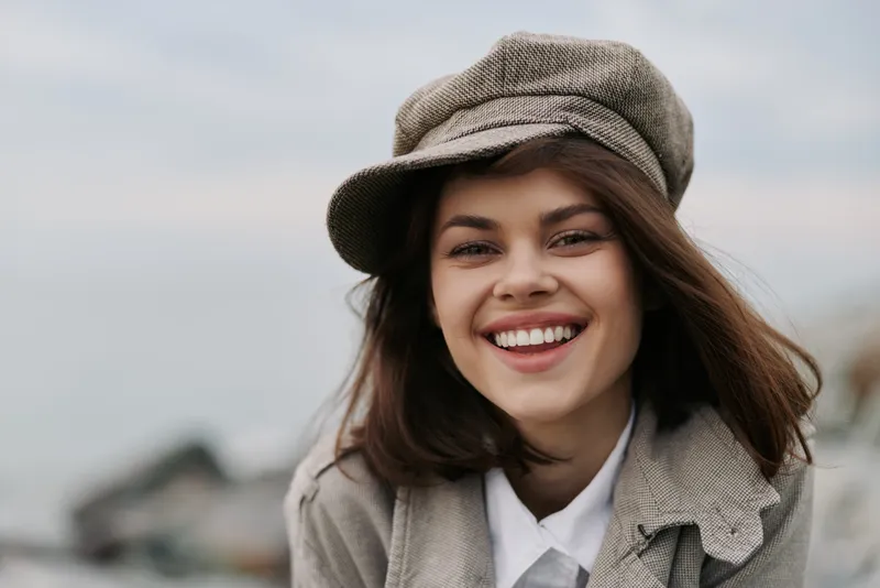 Smiling young woman with long brown hair wearing a grey newsboy cap and coat outdoors. Natural light highlights her happy expression and casual autumn style on a cloudy day.