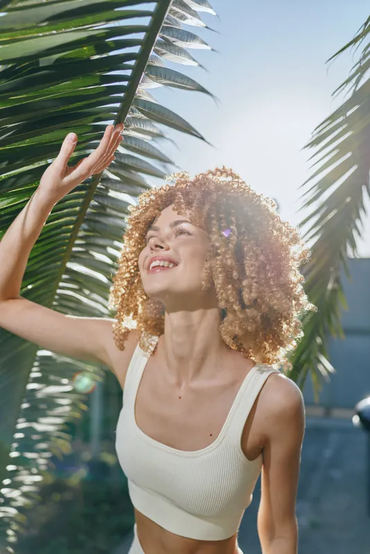 Smiling woman with curly hair enjoys sunny tropical outdoors surrounded by green palm leaves, wearing a white tank top and expressing happiness and freedom.