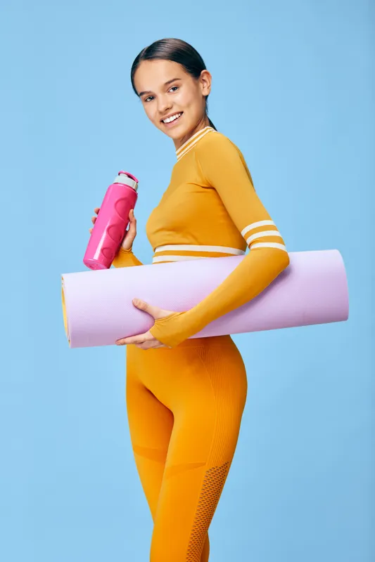 Woman in vibrant orange athleticwear holding a pink water bottle and a rolled yoga mat against a blue studio backdrop, ready for training.