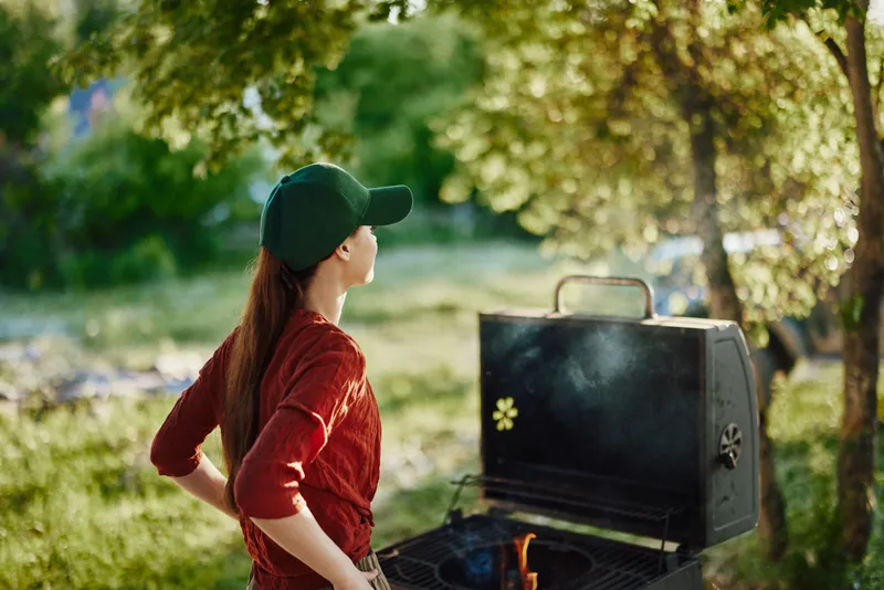 Young woman in casual red outfit and cap, enjoying summer barbecue with grill outdoors, surrounded by greenery, conveying relaxation and joy