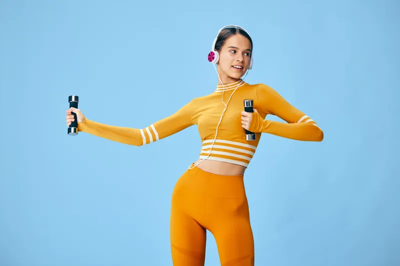 Young woman in orange activewear holds dumbbells and wired headphones in a bright blue studio, striking an energetic workout pose that expresses fitness, sport, strength and healthy lifestyle.
