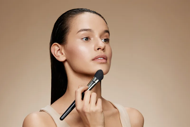 A young model applying makeup with a brush, showcasing a natural beauty look against a soft beige background, reflecting elegance and confidence in beauty and fashion