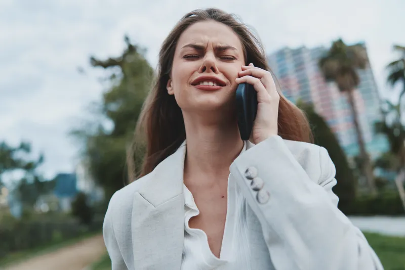 upset woman in white blazer showing frustration during phone call outdoors with blurred city background emotional business communication fall season