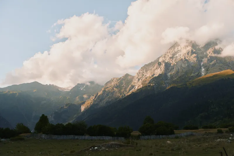 Mountain landscape with rocky peaks and clouds above green forest and grassy field under soft natural light during daytime, peaceful outdoor nature scene with clear sky.