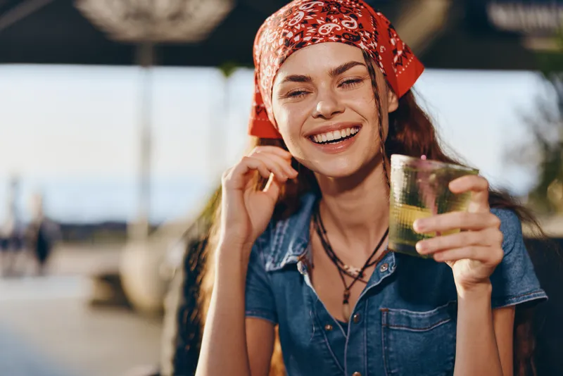 smiling woman holding glass outdoors wearing red bandana and denim shirt enjoying sunny day with blurred background and warm natural light