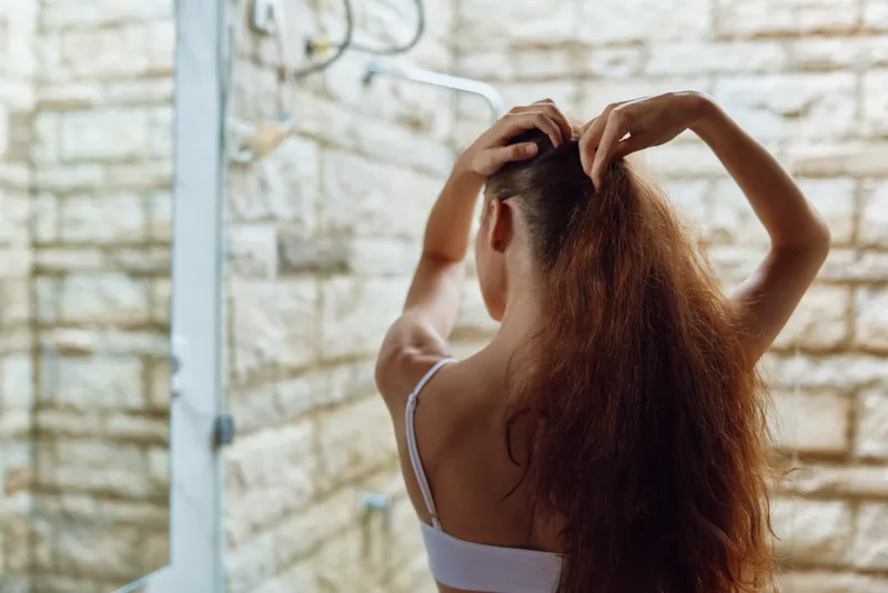 Woman with long curly hair tying ponytail in modern bathroom with stone wall and natural light, showing casual morning routine and self-care moment.