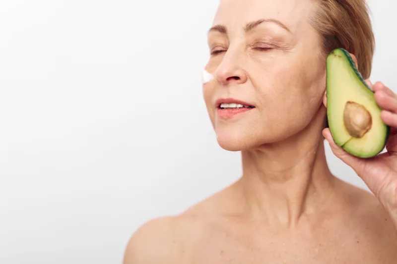 Mature woman with closed eyes holds half of fresh avocado near face with natural cream on cheek. Concept of healthy skin care, natural beauty, and organic facial treatment for aging skin.