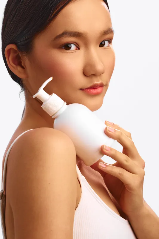 Asian woman holds a white pump bottle in a clean studio setting, smiling softly as she presents skincare routine, calm and confident, conveying a modern beauty moment.