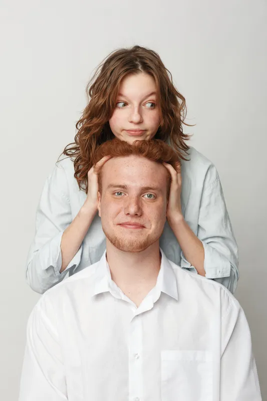 Young man with red hair and beard sits while woman with curly hair stands behind him holding his head. Studio portrait on white background with casual clothing and playful mood.