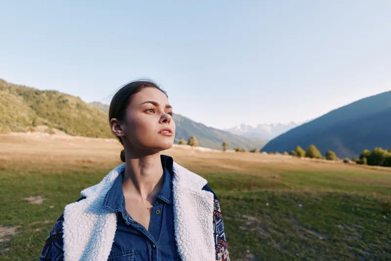 Woman portrait in mountains and meadow, nature travel scene with young female looking thoughtful in denim shirt and sherpa vest, outdoor landscape bathed in soft evening light.