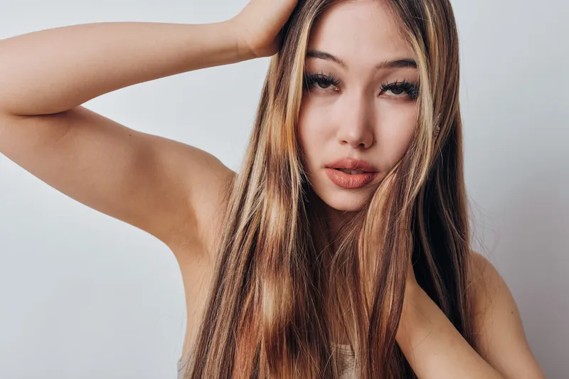 Young woman with long straight hair posing with hand on head against white background, natural makeup and serious expression in studio portrait photo.