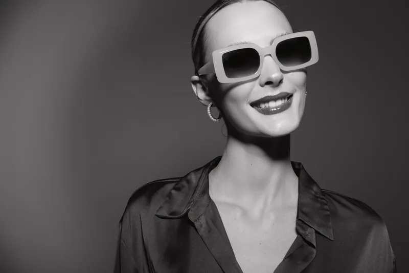 Stylish white woman with fair skin wearing large sunglasses and hoop earrings smiles confidently against a plain background. Concept of fashion, beauty, and modern style.