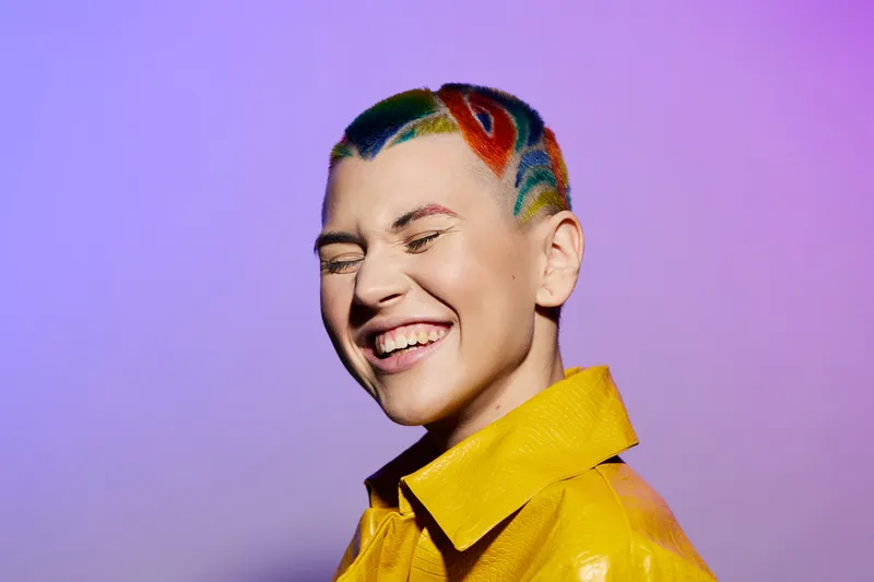 Smiling woman with colorful hair and yellow jacket laughing joyfully against purple background, showcasing vibrant style and upbeat emotion.