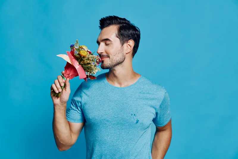 Handsome smiling man wearing a blue t-shirt holding a colorful bouquet of flowers close to his face against a bright blue background. Celebration concept.