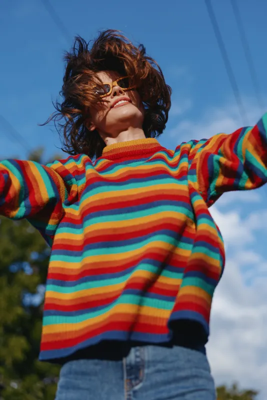 Woman in her mid 20s wearing a colorful striped sweater and sunglasses, jumping with arms out and smiling outdoors under bright blue sky in casual outfit and denim jeans, carefree.