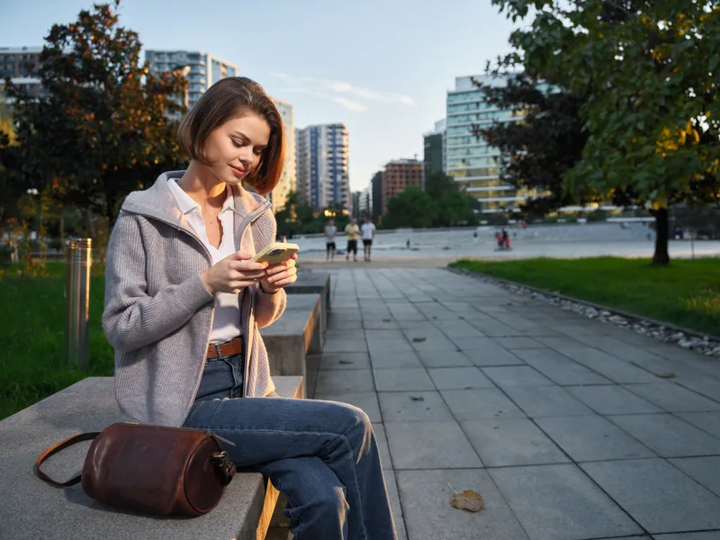 A cheerful professional woman sits on an outdoor bench, texting on her smartphone in a modern urban park. She smiles warmly, appears engaged, and exudes a confident, positive business vibe.