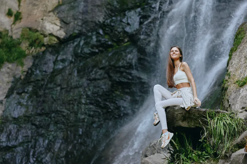 Young woman sitting on rock near waterfall in sporty white outfit, smiling and enjoying nature, surrounded by lush greenery and rocky cliff background.