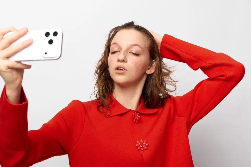 Young woman in red sweater takes a selfie with smartphone, eyes closed and hand in hair. Studio portrait on white background, concept of self-expression and casual lifestyle.