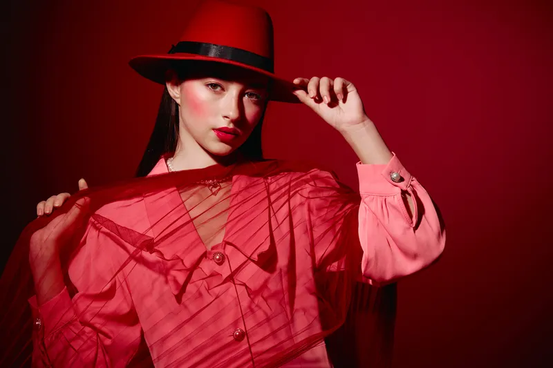Fashion model in a red fedora hat poses holding beret. She wears a red sheer outfit with dramatic makeup against a deep red background. Creative beauty studio shoot.