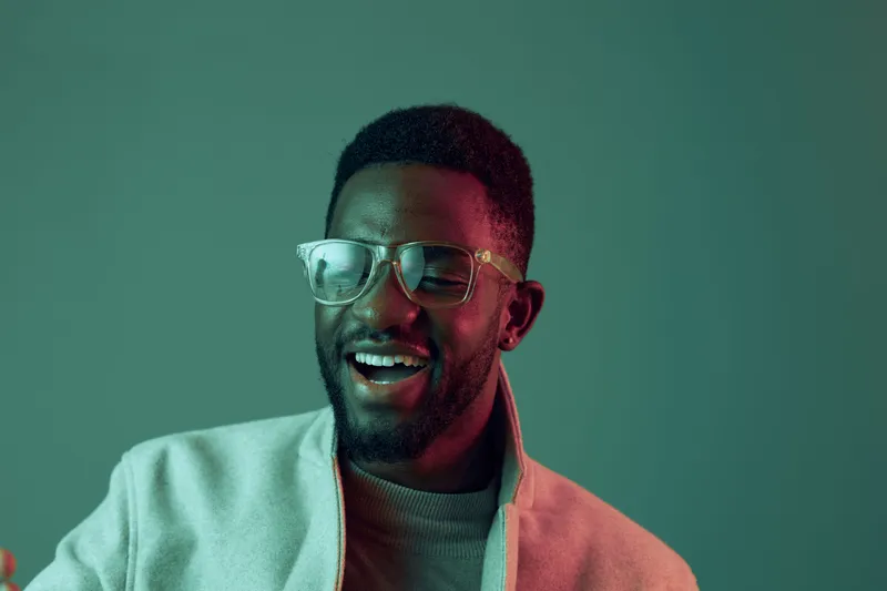 Happy young African man in glasses smiling against a modern green background, wearing a stylish jacket, perfect for lifestyle and fashion concepts