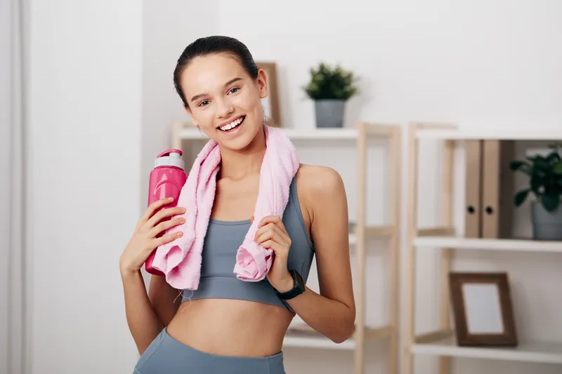 A fit woman in athletic wear smiles, holding a pink water bottle with a towel draped over her shoulders in a bright indoor studio, conveying energy, health, and a ready-for-workout vibe.