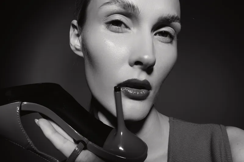 Close-up portrait of a fair skinned Caucasian woman holding a high heel shoe near her face. Concept of fashion, elegance, beauty, and stylish footwear in monochrome studio lighting.