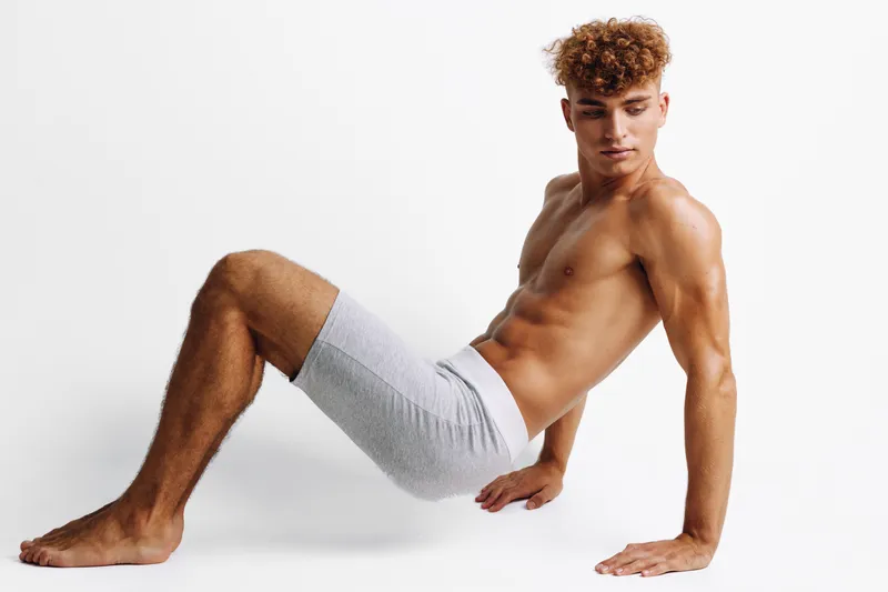 Young athletic man with muscular body posing shirtless in gray shorts on white background for fitness, health, and workout concept with toned skin and curly hair.
