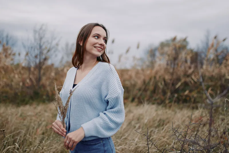 A young woman in a cozy light blue sweater stands in tall grass, holding dried reeds and smiling softly; an authentic, candid portrait capturing natural emotion and realism.