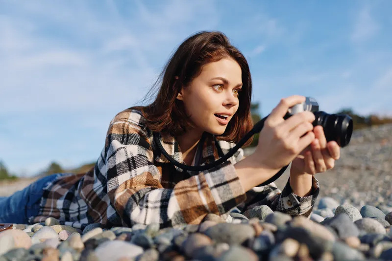 Woman lies on pebbles outdoors, holding a camera for photography on the shore, wearing plaid shirt, natural light, focused expression, relaxed outdoor mood and creative hobby.