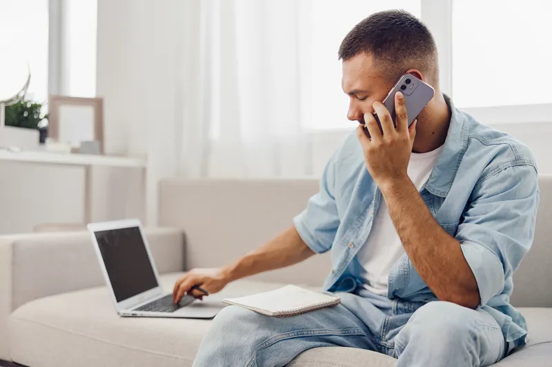 A focused man engaged in a phone call while working on a laptop, conveying productivity and professionalism in a bright modern workspace. Ideal for remote work themes.