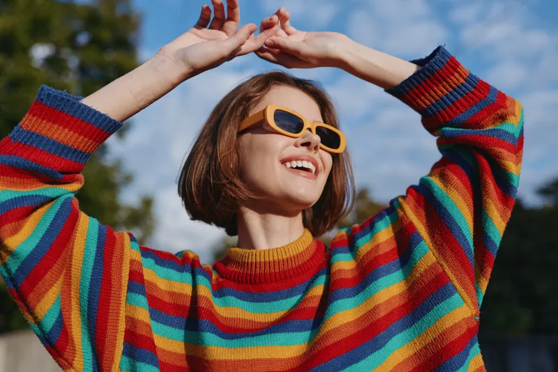 woman in her twenties wearing a colorful striped sweater and yellow sunglasses, smiling and raising hands outdoors in a sunny park, joyful casual outfit lifestyle portrait