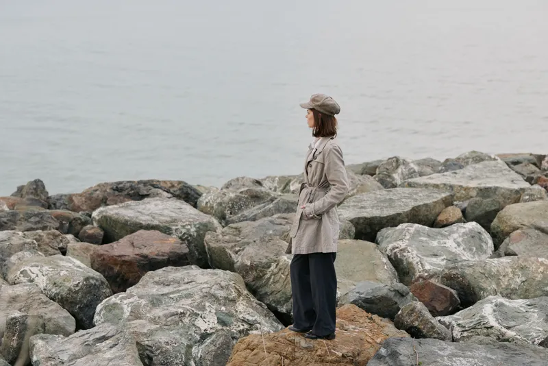 Woman in beige coat and hat stands on rocky shore looking at calm sea. Overcast sky and casual clothing create a peaceful, contemplative outdoor mood by the water.