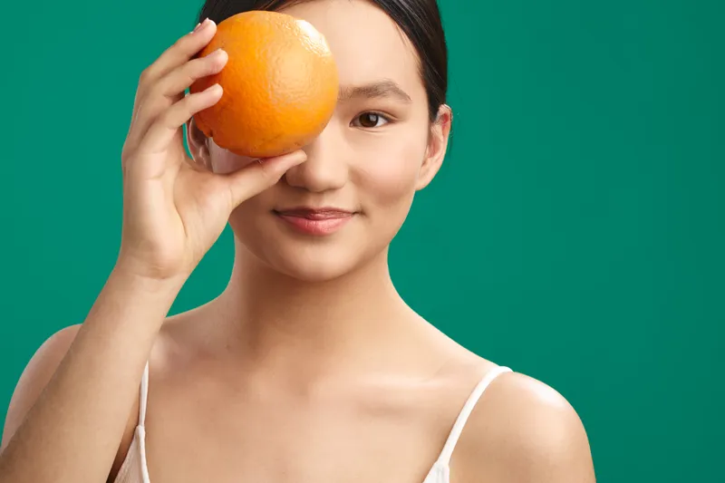 asian woman holds an orange to her eye in a playful portrait, her calm smile radiates confidence and creativity, against a vivid green background that enhances a fresh, modern mood.