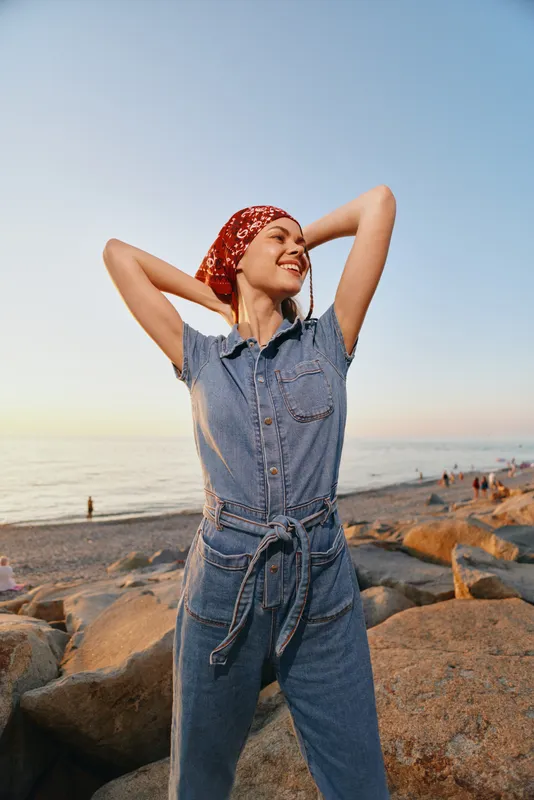 Lifestyle woman in modern boho-western denim street style wearing a red bandana, smiling and relaxing on a rocky beach at sunset with film color tones.