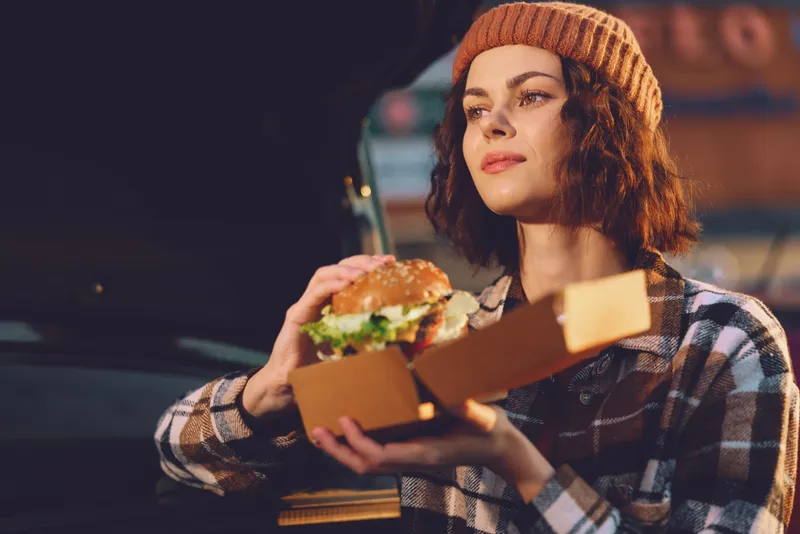 Woman holding burger in takeout box at evening urban street, candid lifestyle portrait with beanie and relaxed expression, authenticity, golden hour glow and emotional storytelling.