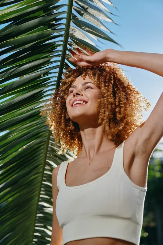 smiling woman with curly hair enjoys sunny day outdoors, wearing white tank top, surrounded by green palm leaves under blue sky