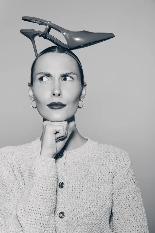 Fair skinned caucasian woman with thoughtful expression balances high heel shoe on head, wears sweater and earrings.