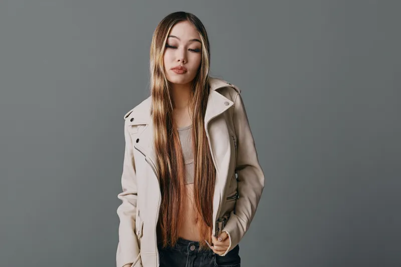 Young woman with long braided hair wearing beige jacket and top standing against gray background with closed eyes and relaxed expression in studio portrait.