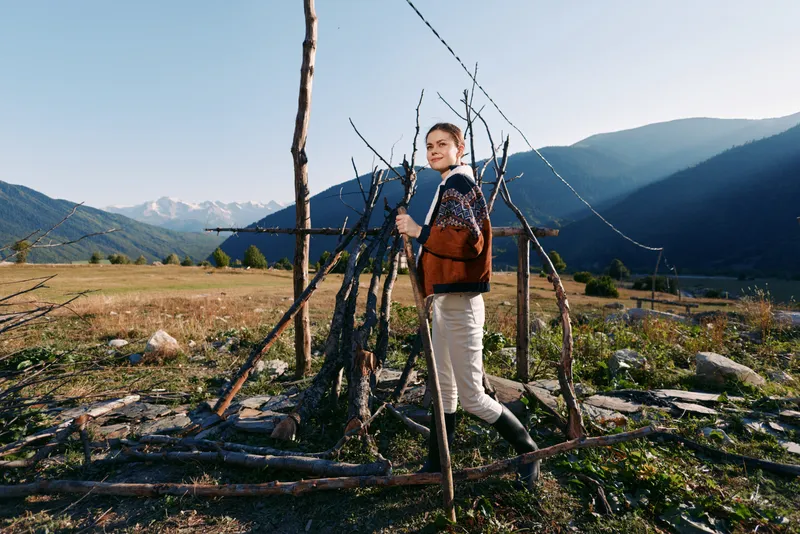 Woman with backpack standing in a wooden makeshift shelter in a field, mountains and blue sky in background. Nature scene with meadow, rustic sticks, smile and relaxed outdoor travel.