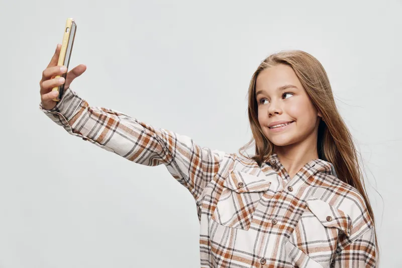 Girl with long hair wearing plaid shirt smiling and holding smartphone for selfie on white background. Child, technology, casual style with happy expression and mobile device in hand.