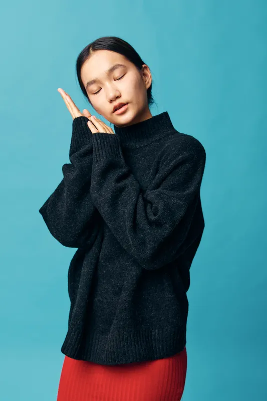Young woman wearing black sweater and red skirt with eyes closed and hands together near face posing like sleeping against blue background in studio portrait.