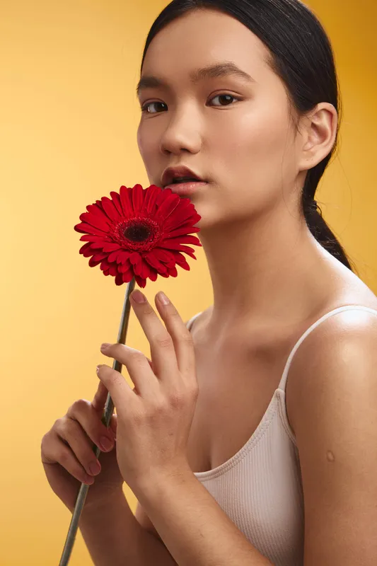 Asian woman gazes softly at the camera while holding a vibrant red flower against a warm yellow backdrop, conveying elegance, poise, and a contemplative studio portrait mood.