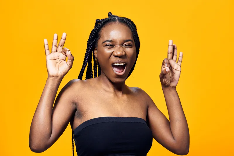 Young Black woman with braided hair expressing joy against a vibrant yellow background, showcasing confidence and happiness in a modern style